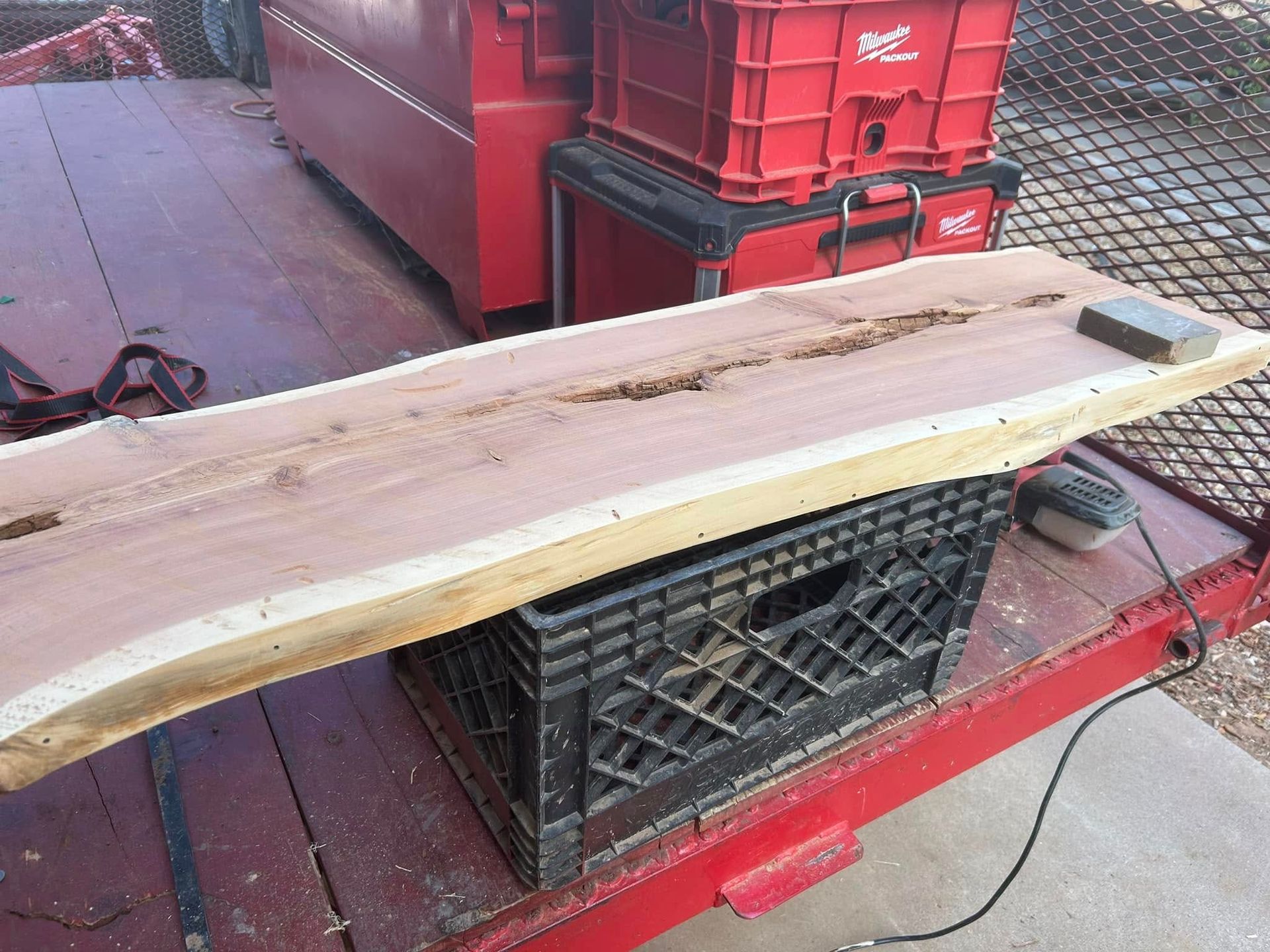 A wooden slab with a live edge rests on a black crate. A red trailer and tools are in the background.
