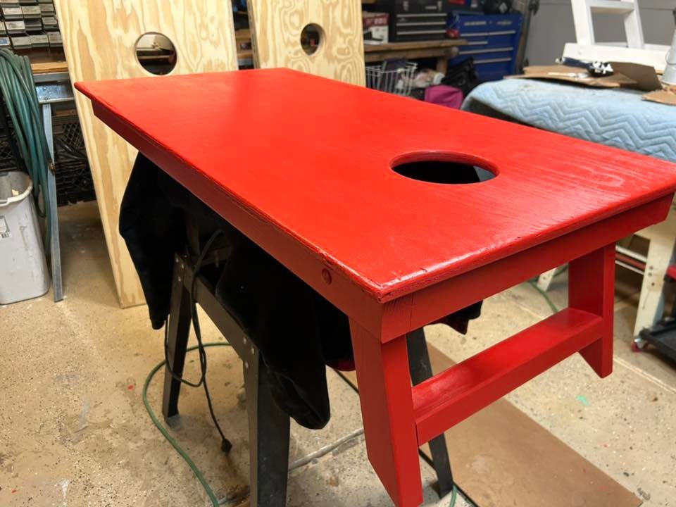 Red painted cornhole board with hole, resting on a sawhorse. Another board sits in the background.