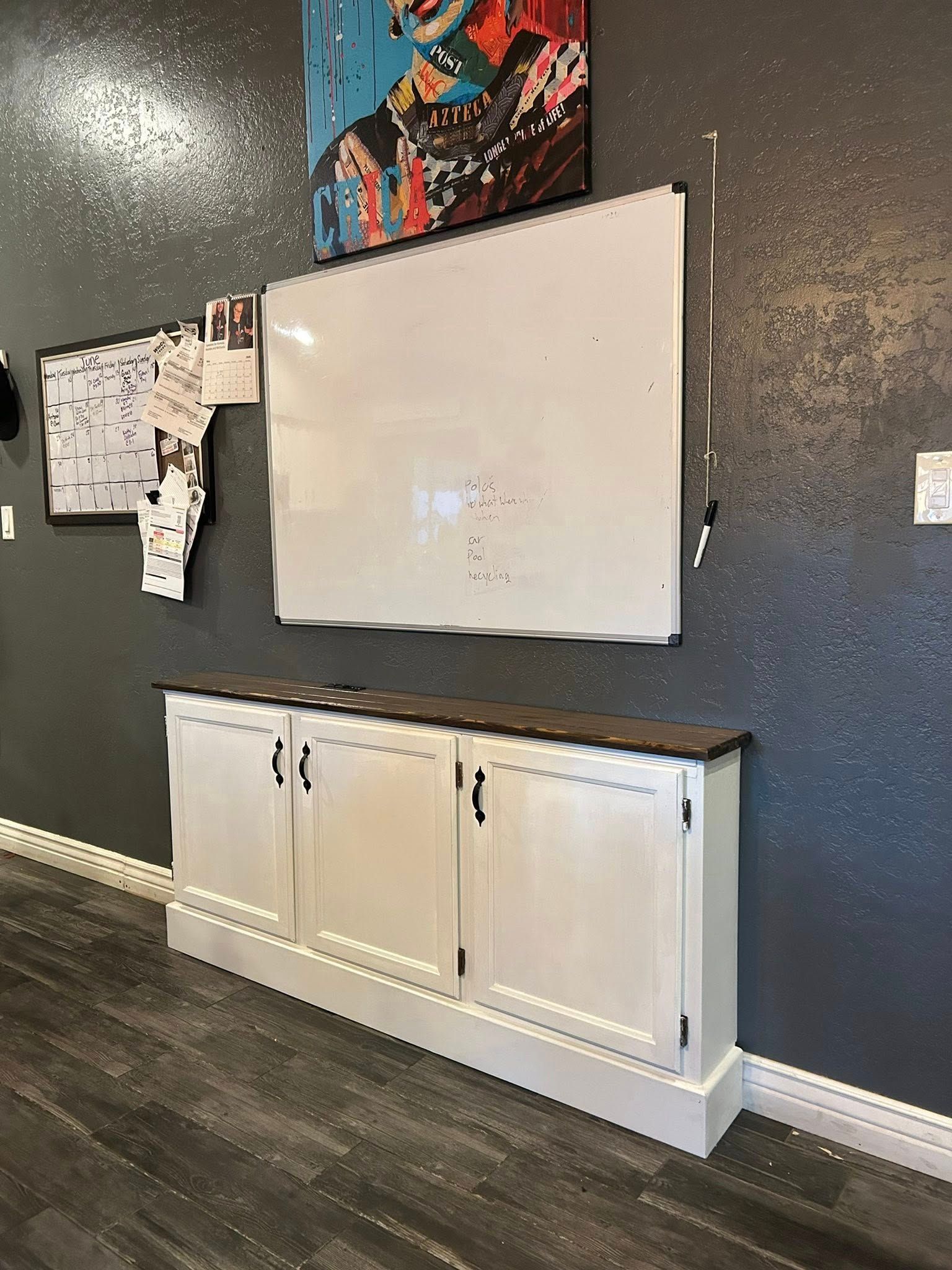 Whiteboard and cabinet on a gray wall, with artwork above, in a room with dark wood flooring.