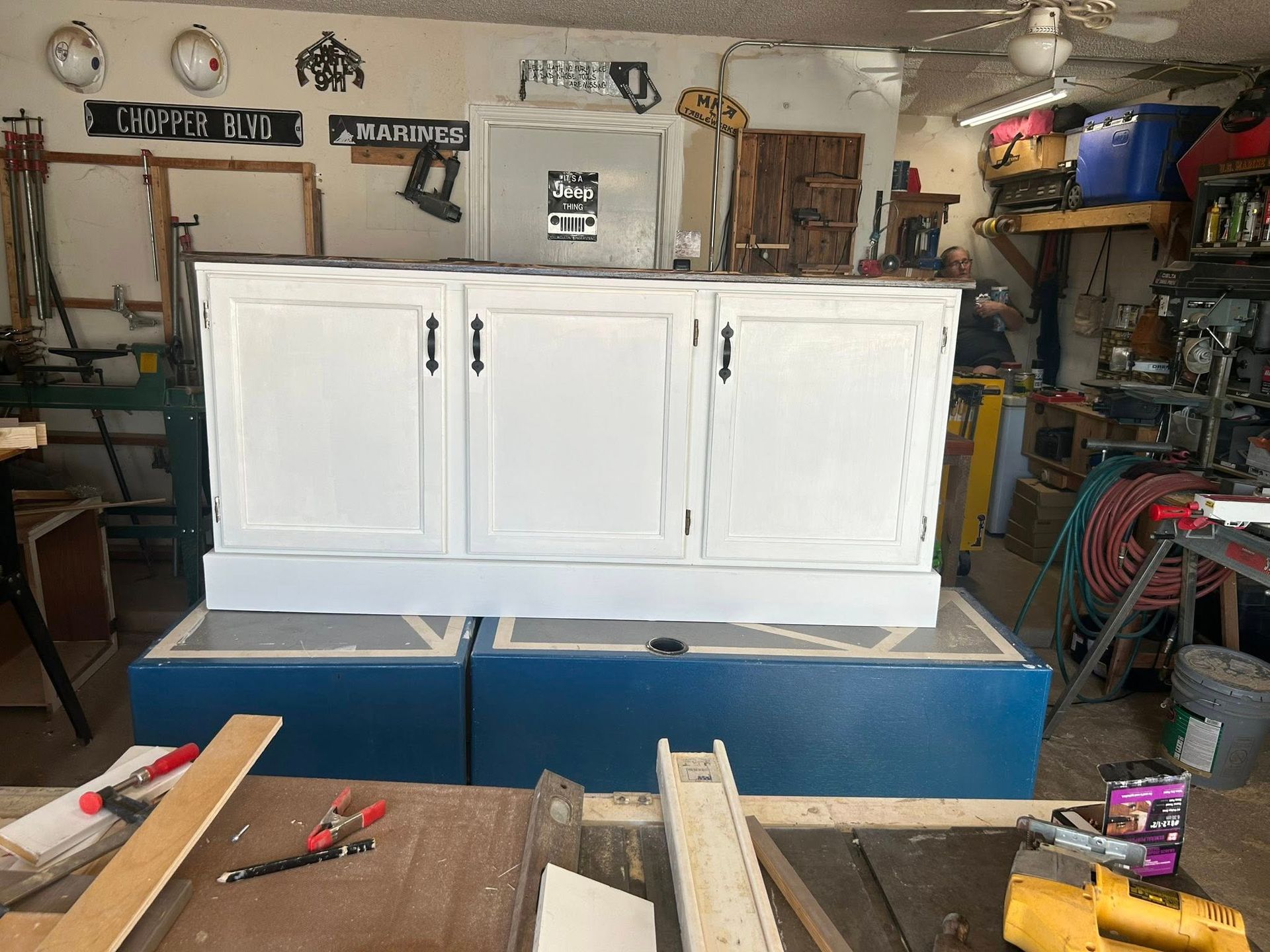 White cabinet with three doors sits on two blue boxes in a workshop setting.
