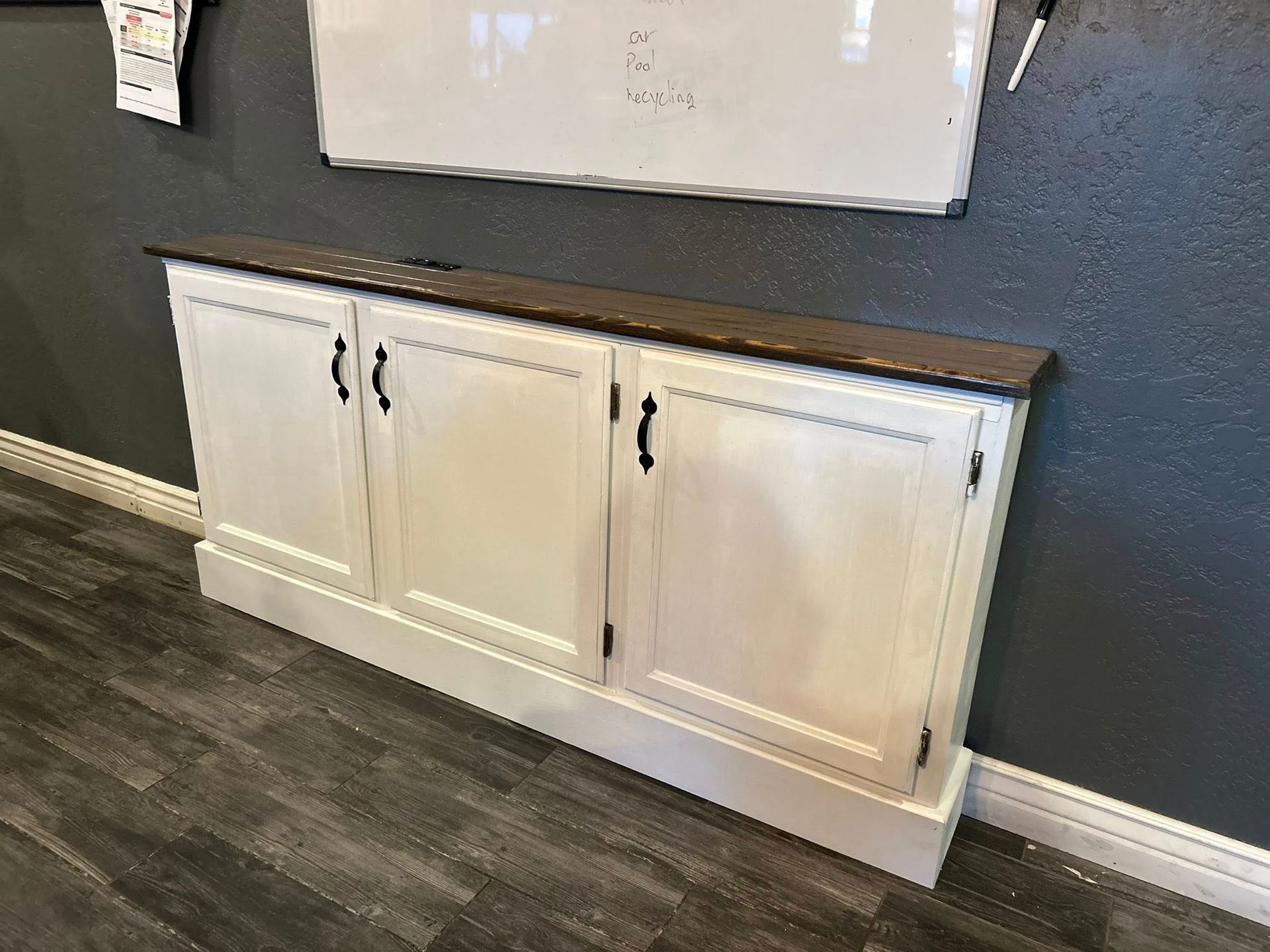 White cabinet with black hardware and a brown top, against a gray wall.