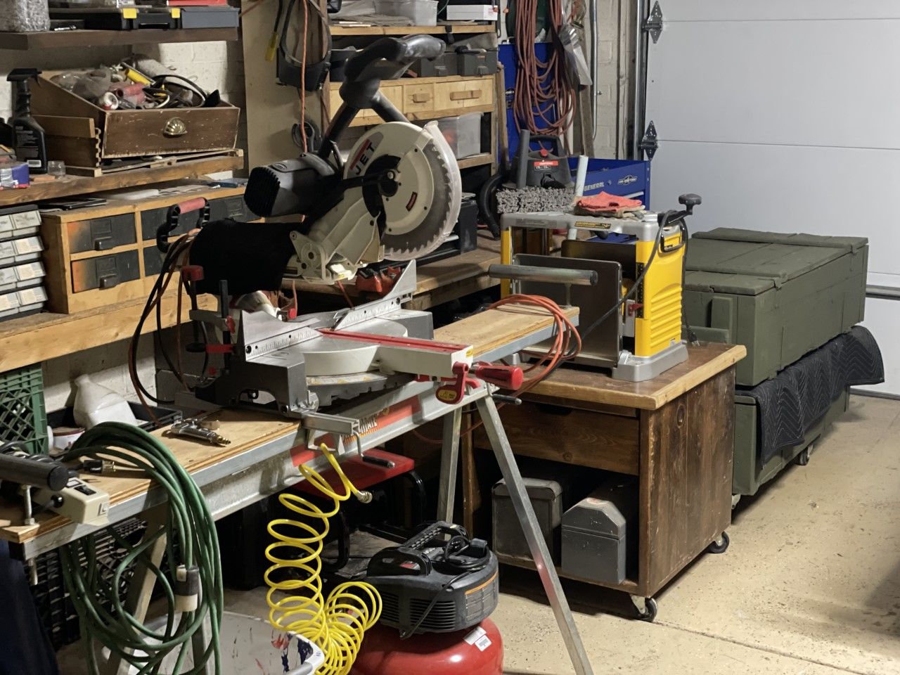 A cluttered workshop with a miter saw, planer, workbench, and air compressor.