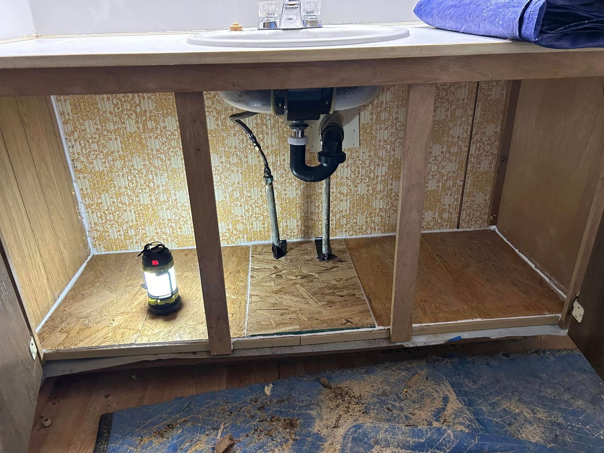 Interior of an open bathroom vanity cabinet under a sink, with exposed plumbing and a work light.