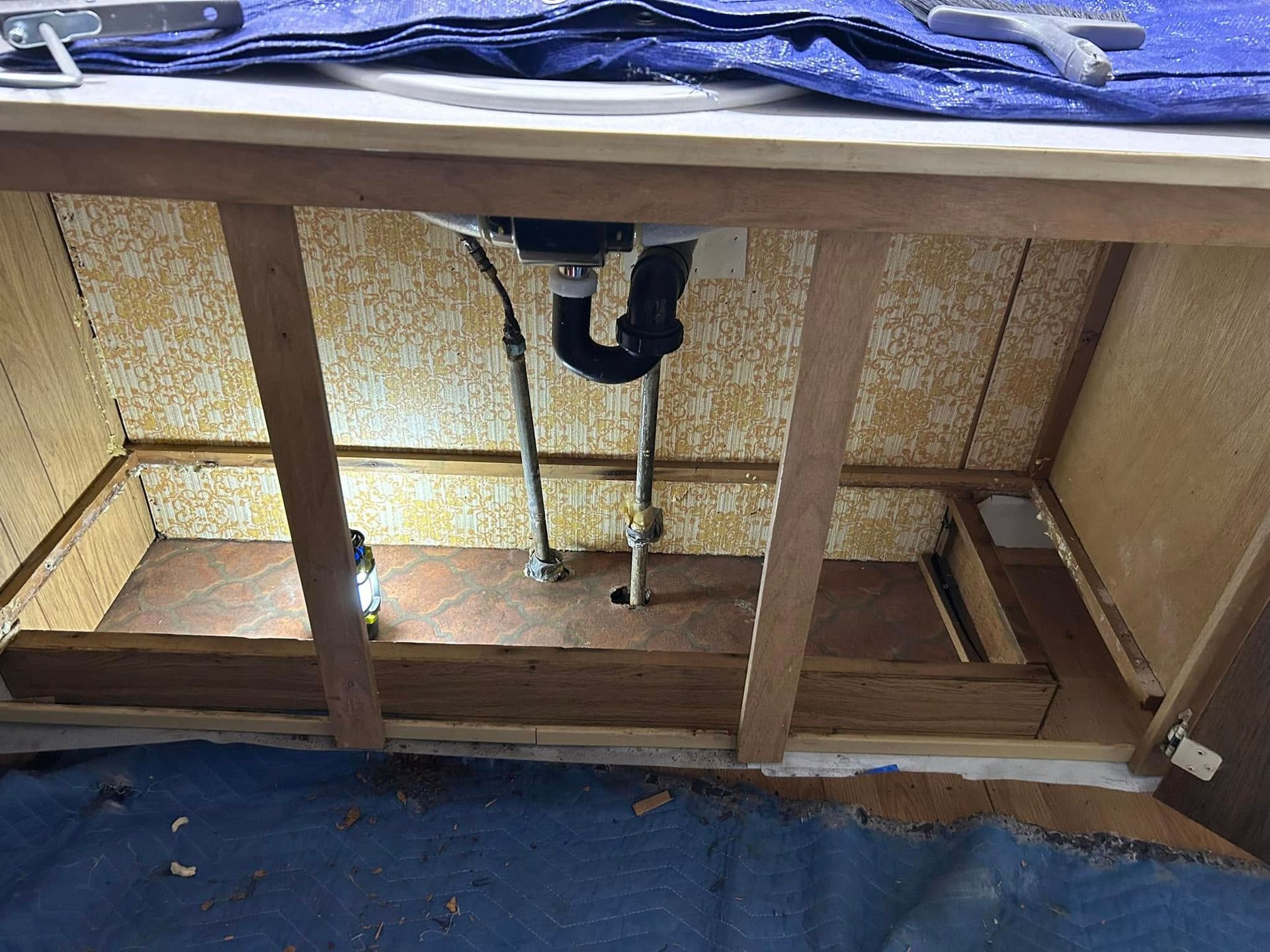 Inside view of a cabinet under a sink with exposed plumbing and wooden support beams. Brown, yellow, and metal colors.