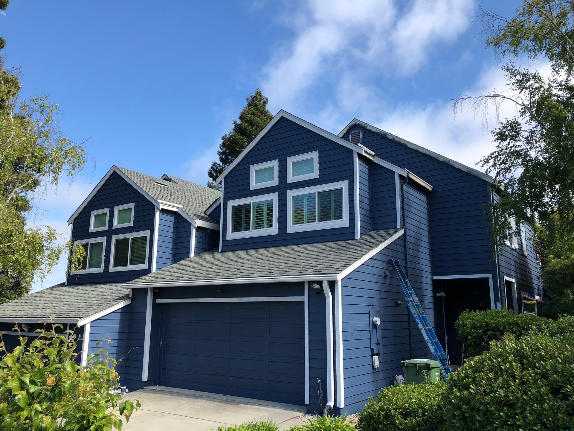 Blue two-story house with gray roof and white trim against a partly cloudy blue sky.