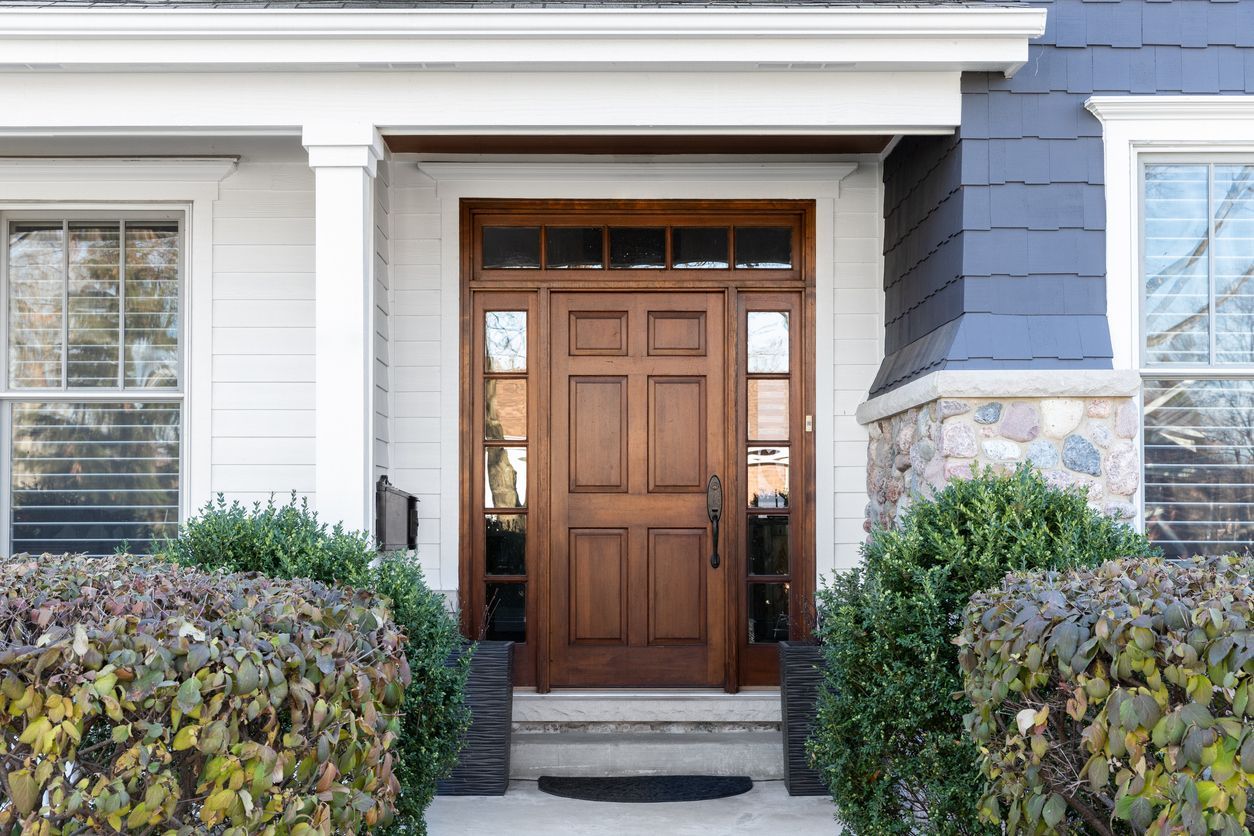 Wooden front door with sidelights, under a white portico, with shrubs on either side.