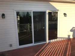 Sliding glass door on a deck, framed by white siding, two outdoor sconces. Sunlight casts long shadows.