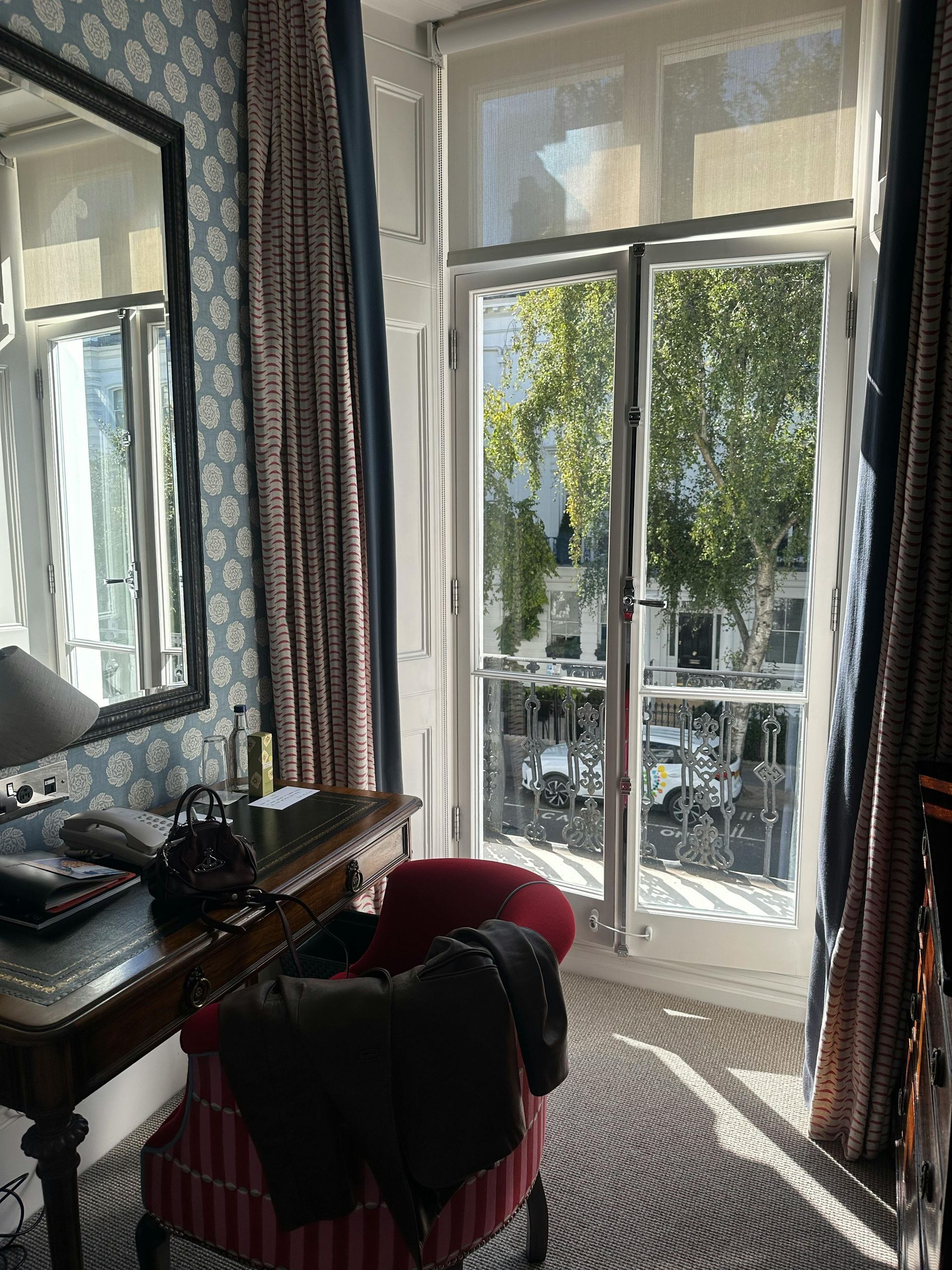 Interior view of a room with a desk, chair, mirror, and a window overlooking a street with trees.