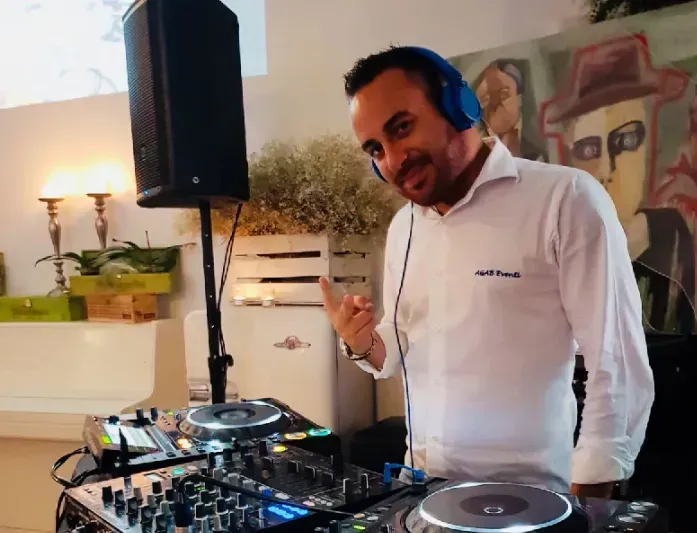 Un DJ con cuffie blu, seduto dietro un mixer, sorride in un luminoso locale al chiuso.