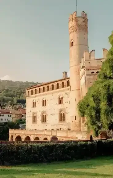 Castello in pietra con finestre ad arco e torre rotonda accanto a un prato verde e a una collina