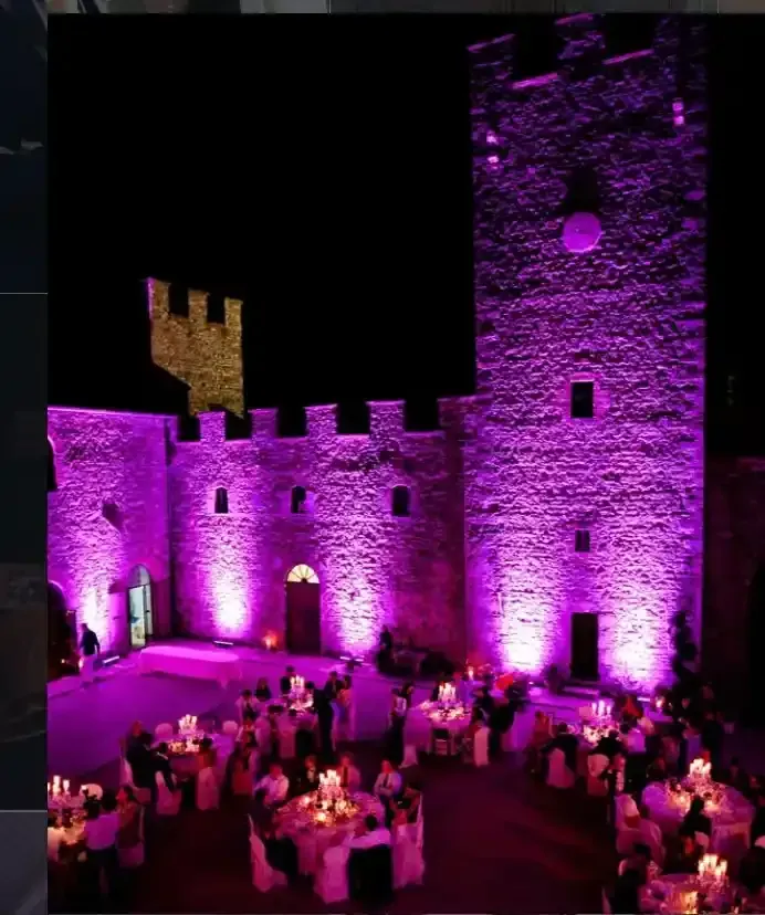 Cortile del castello in pietra illuminato di viola, con gli ospiti che cenano a tavoli rotondi sotto il cielo notturno.