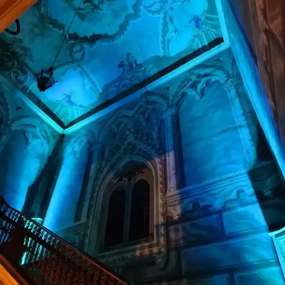 Interno sfarzoso illuminato di blu con soffitto dipinto, finestra ad arco e scalinata in una grande sala