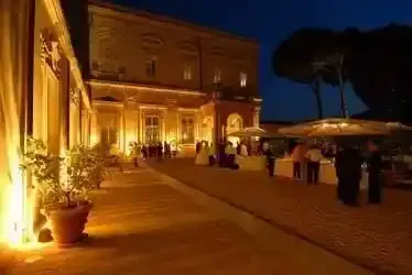 Cortile illuminato di un edificio storico al crepuscolo, con persone riunite su una terrazza lastricata.