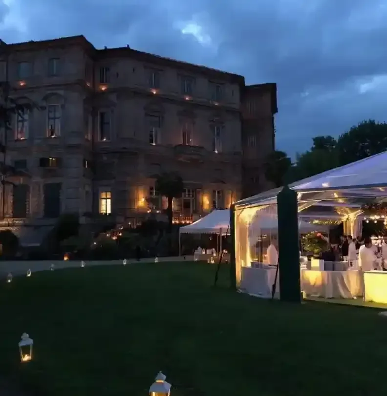 Evento serale all'aperto accanto a una villa illuminata, con un tendone bianco sul prato sotto un cielo azzurro nuvoloso.