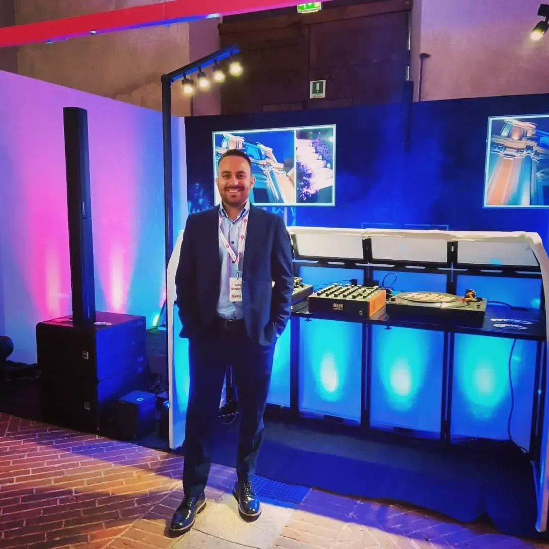 Un uomo in piedi davanti a uno stand fieristico illuminato di blu, con schermi e tavoli espositivi sullo sfondo.