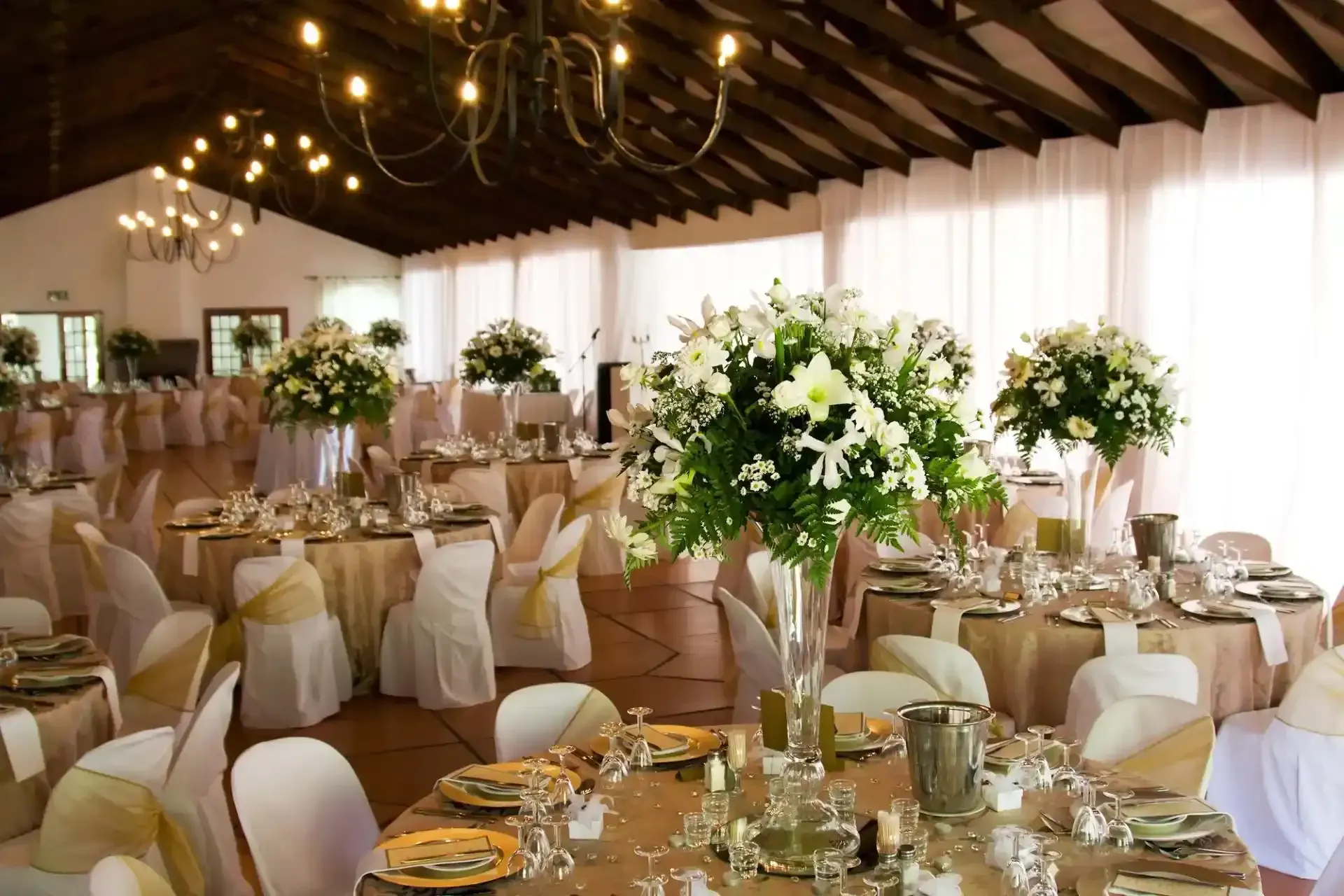 Elegante sala ricevimenti di nozze con tavoli rotondi, sedie bianche e grandi centrotavola floreali illuminati da luci calde.