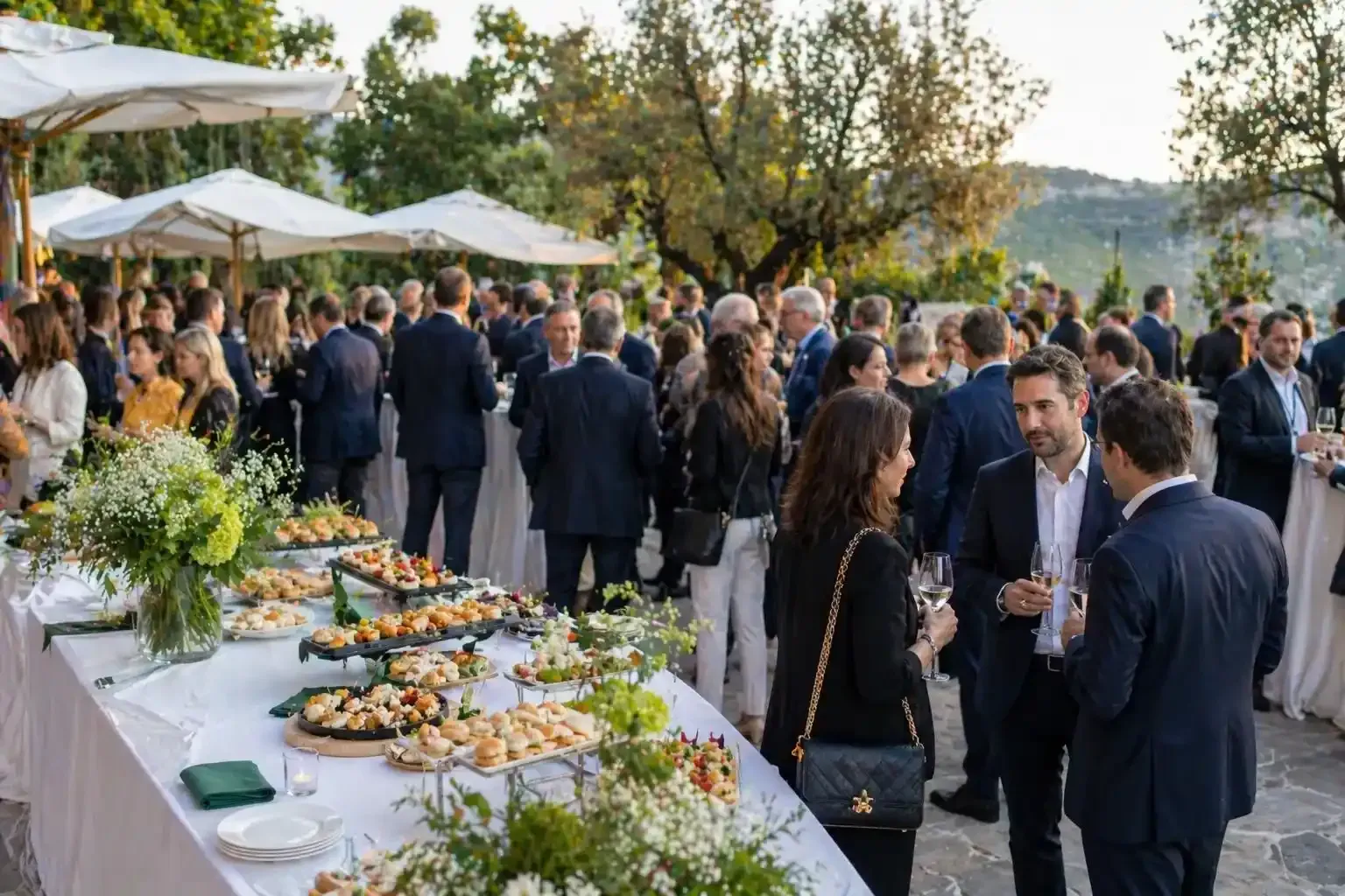 Ricevimento con cocktail all'aperto, con gli ospiti che socializzano attorno a un tavolo da buffet sotto ombrelloni bianchi al tramonto.