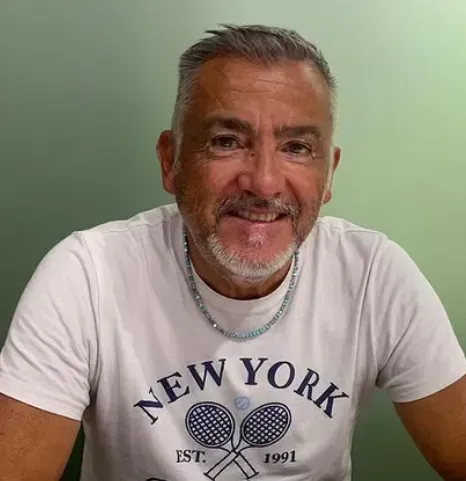 Un uomo sorridente, con indosso una maglietta bianca con la scritta "New York", è seduto contro un muro verde chiaro.