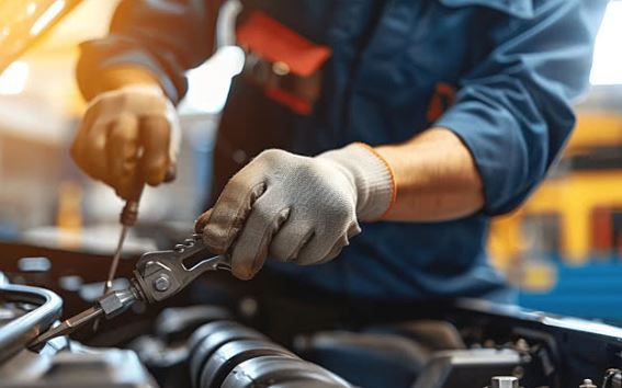 Mechanic in blue overalls using tools to work on a car engine, wearing grey gloves, in a well-lit shop.