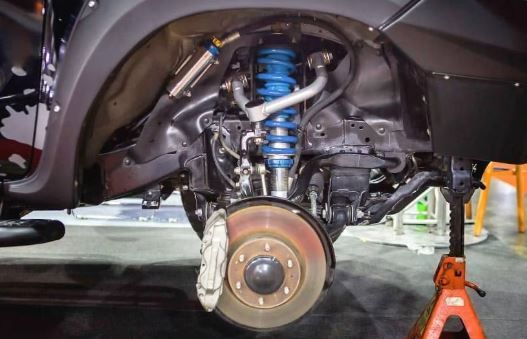 Truck's front suspension system detailed up close, showcasing blue coil springs, shocks, and brake components. The vehicle is raised on a jack stand.