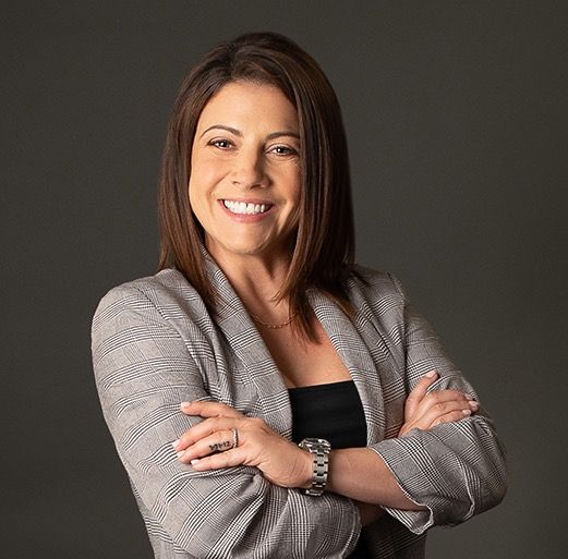 A woman in a gray jacket is smiling with her arms crossed.