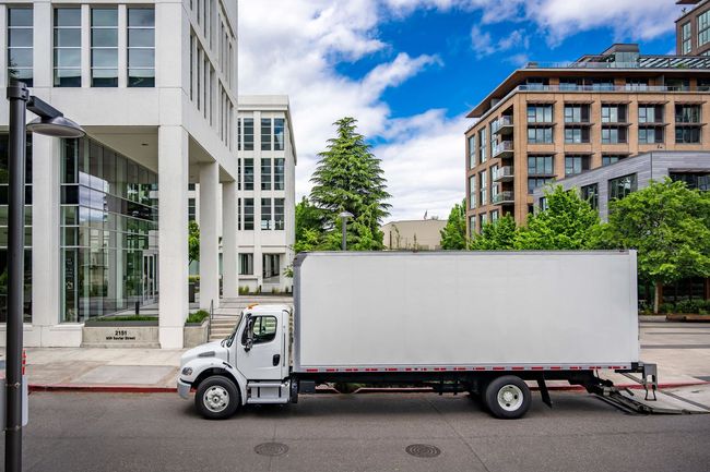 A white moving truck is parked on the side of the road in front of a building.