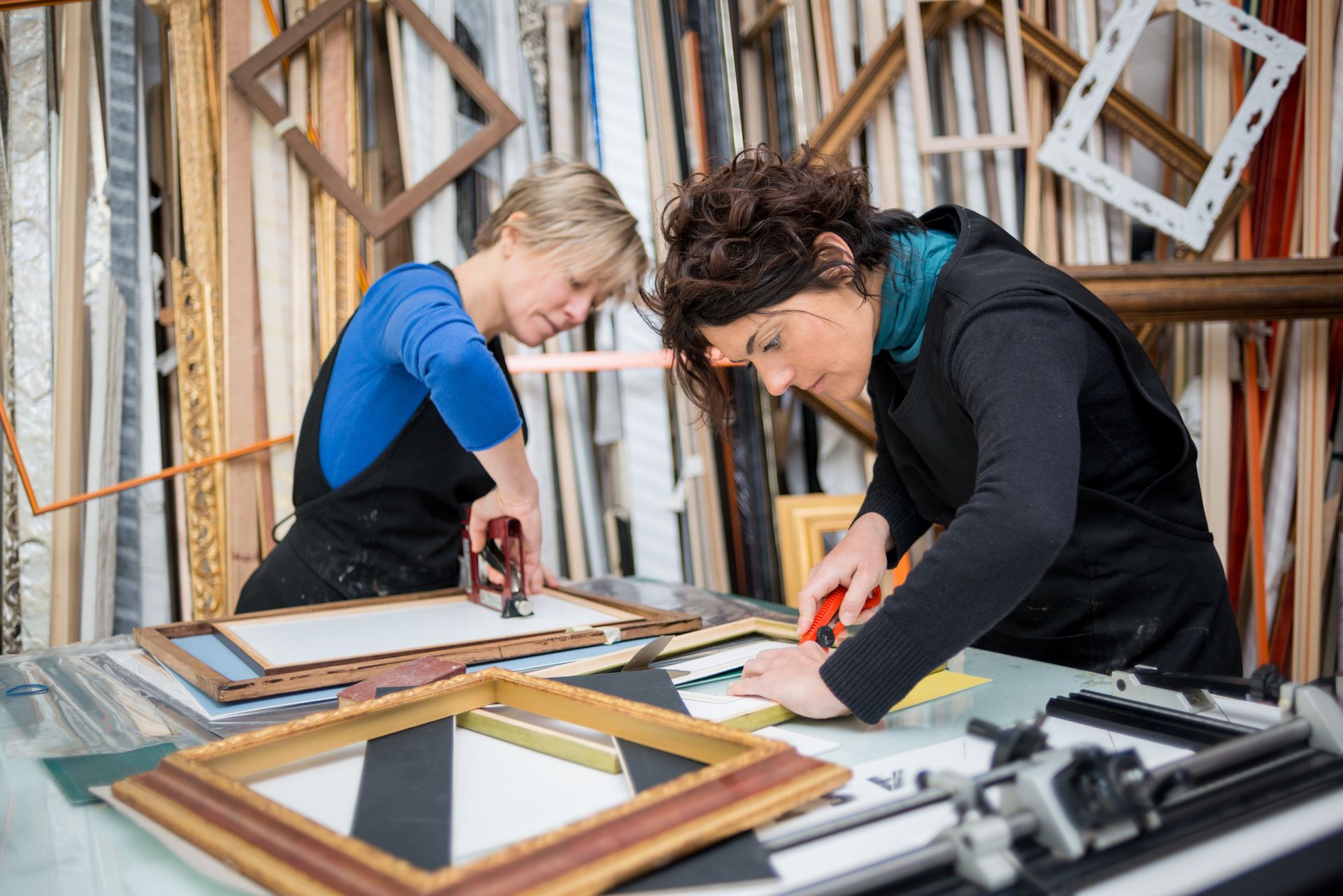 Two craftswomen prepare picture frames on top of a table, with more frames in the background.