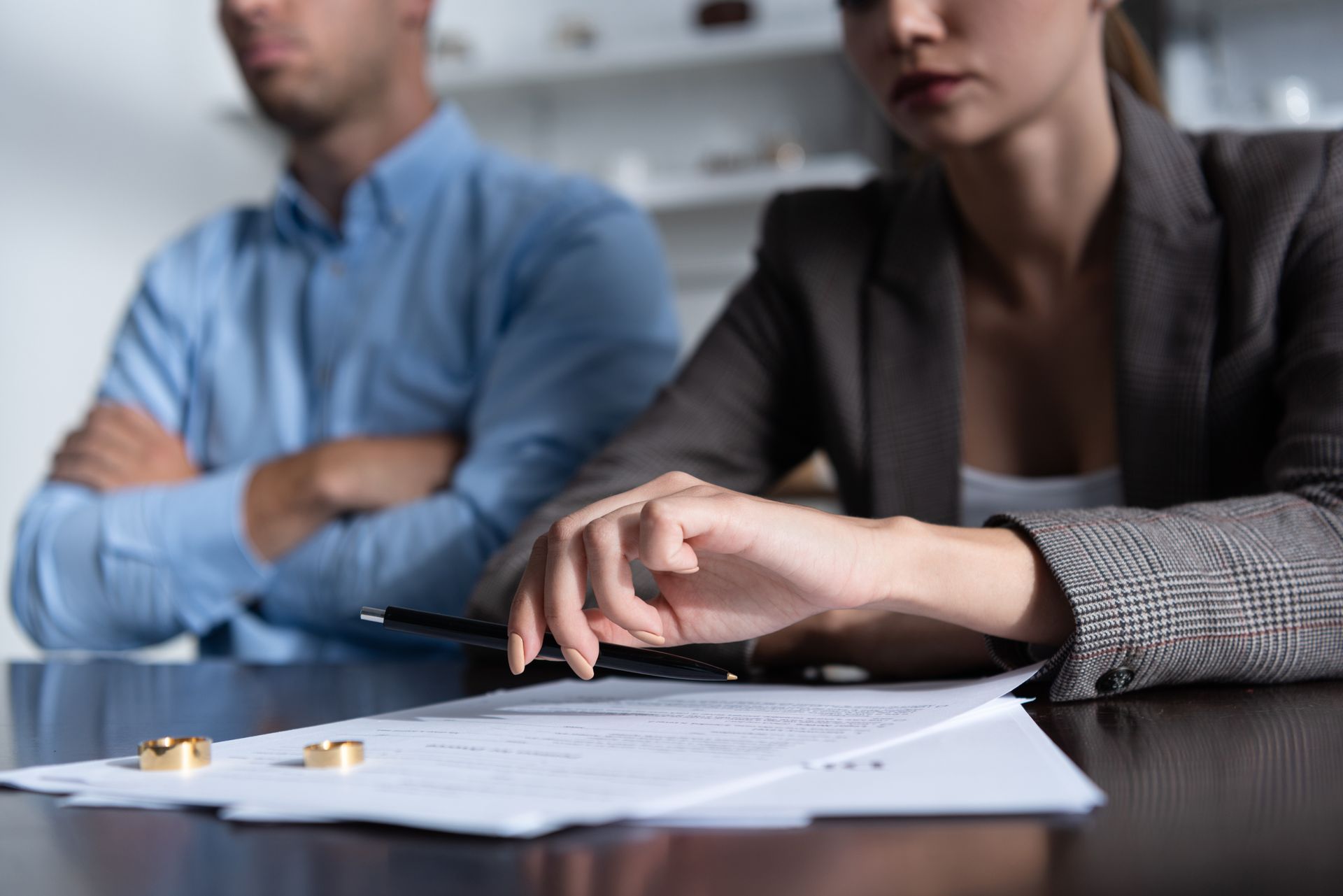 A man and woman sit at a table with two wedding rings resting on divorce papers, appearing somber and distant.