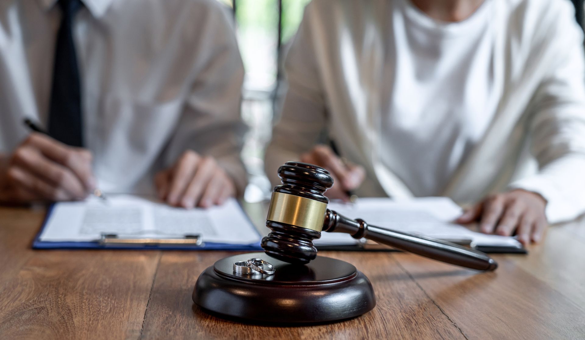 A judge’s gavel and two wedding rings resting on a wooden table in front of people signing legal divorce documents.