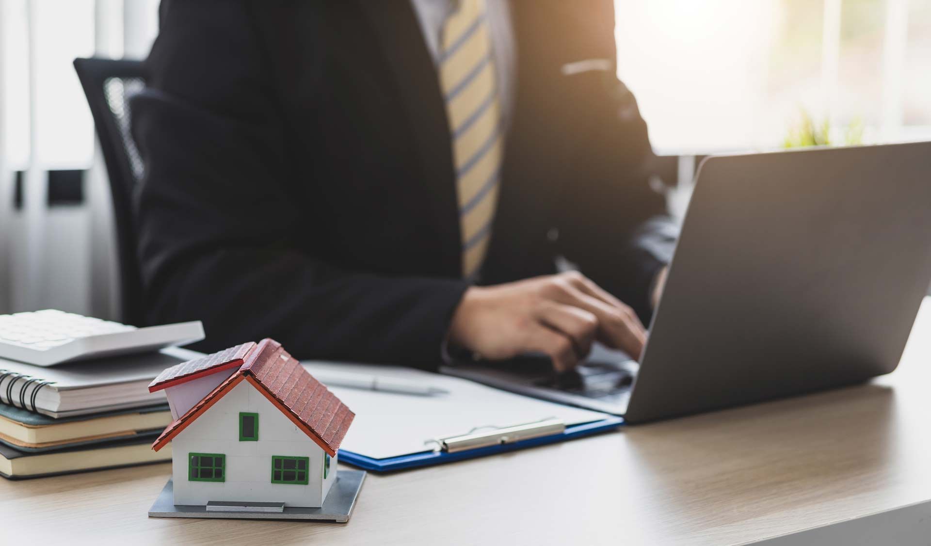 A person in a suit works on a laptop at a desk featuring a small house model and a stack of notebooks.