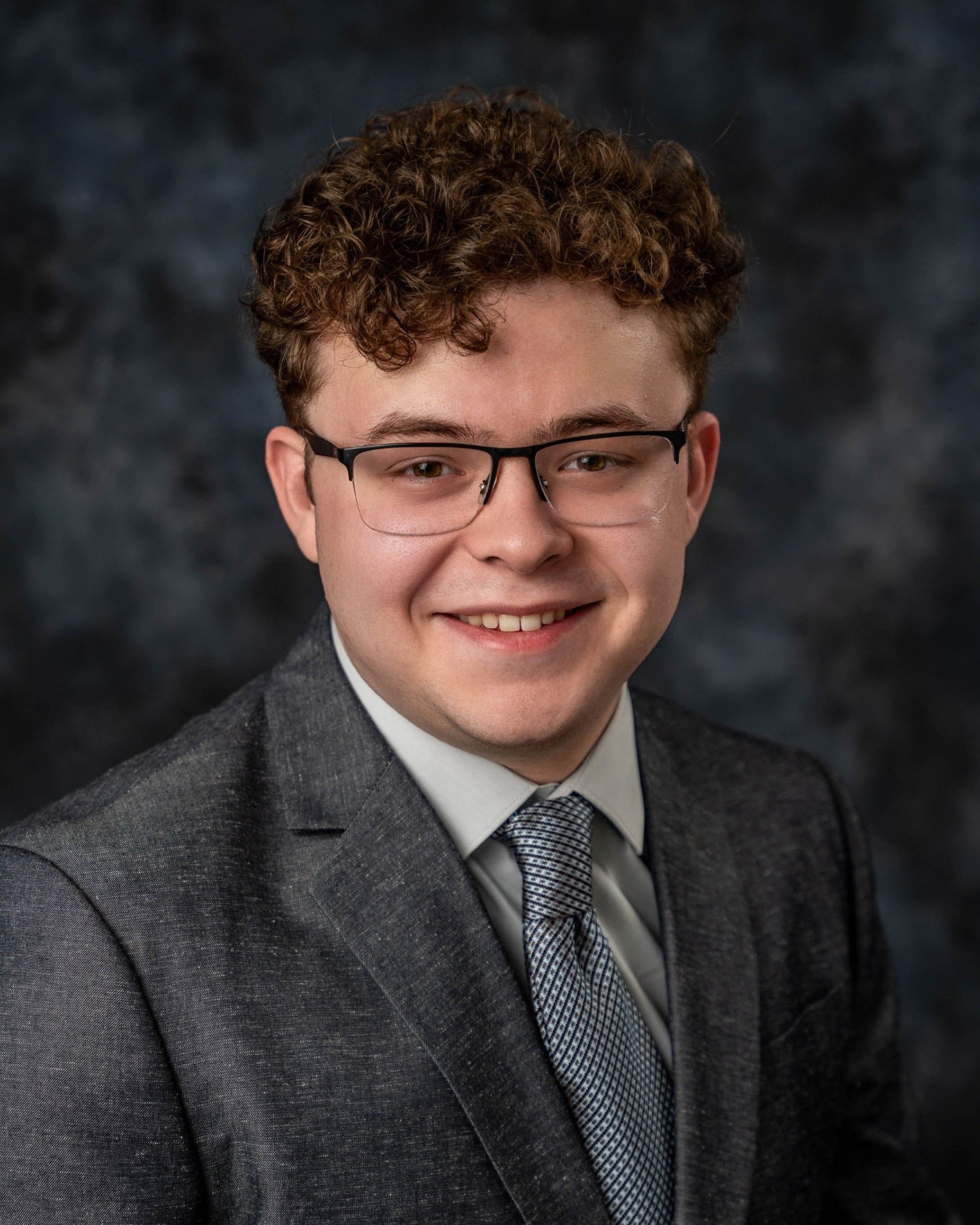 Young man in glasses, blazer, and tie smiles at the camera, posing for a portrait.