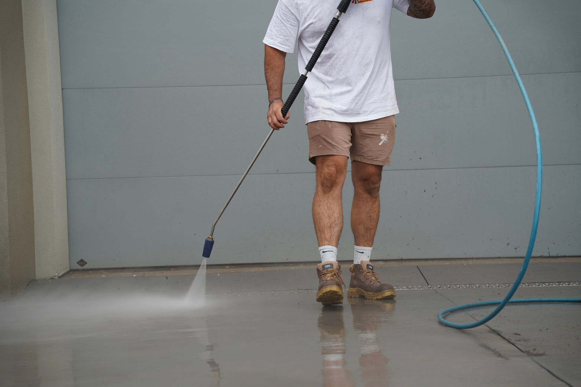Person power washing a concrete surface with blue hose in front of a closed garage door.