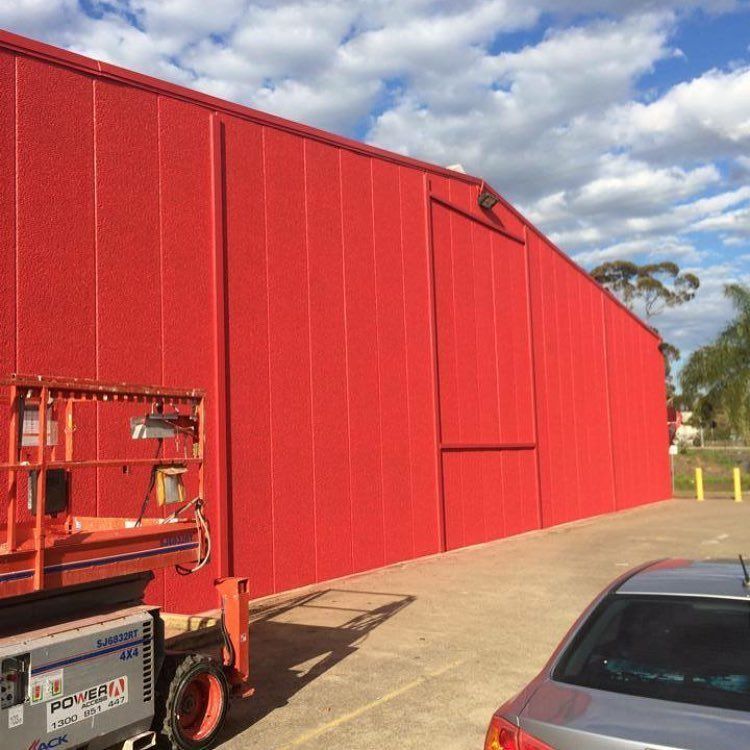 Red industrial building with a large door, an orange lift, and a car parked on concrete.