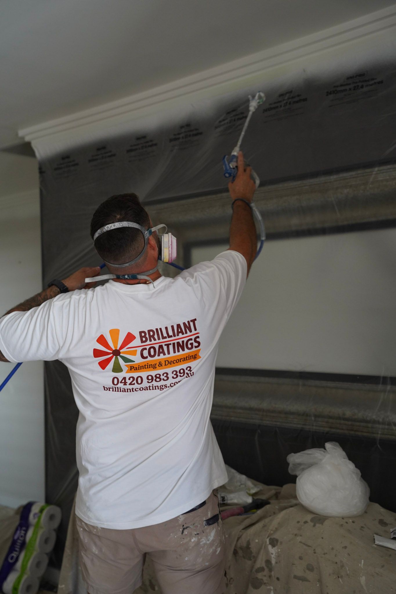 Man painting crown molding with a sprayer. White shirt, protective mask, and dark paint on the wall.