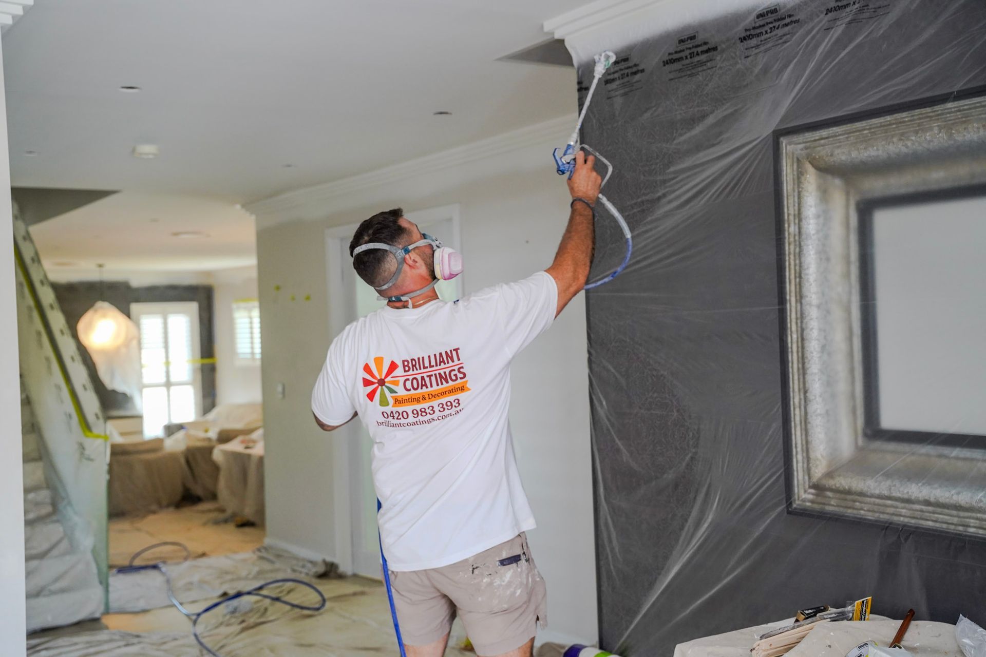 Person sprays paint on molding with a respirator. Interior hallway with plastic sheeting covering walls.