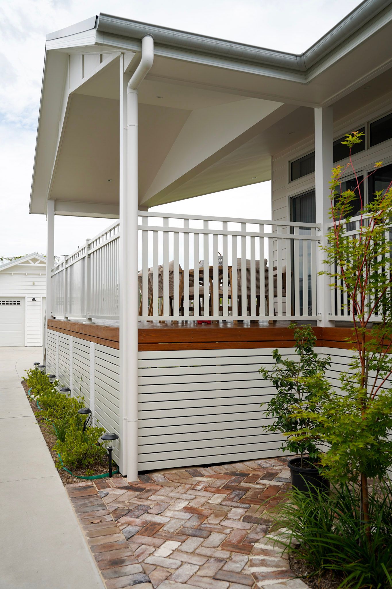 White porch with railing and latticework, wooden deck, brick pathway, green plants, and a partial view of a white building.