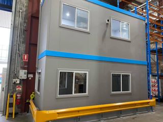 Two-story gray modular building with blue trim and yellow safety barriers, set in a warehouse.