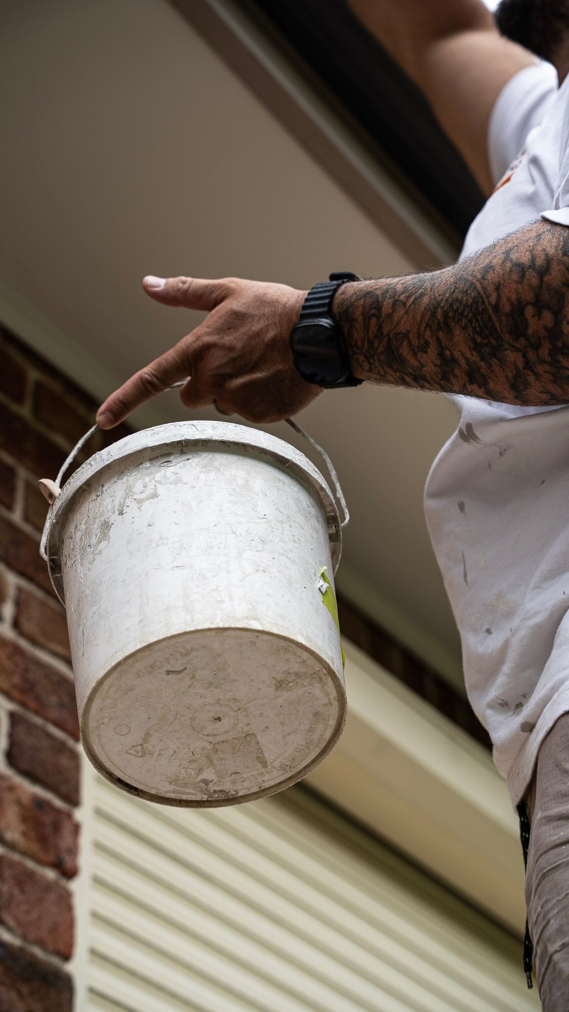 Person holding a white paint bucket near a brick wall, pointing at it.