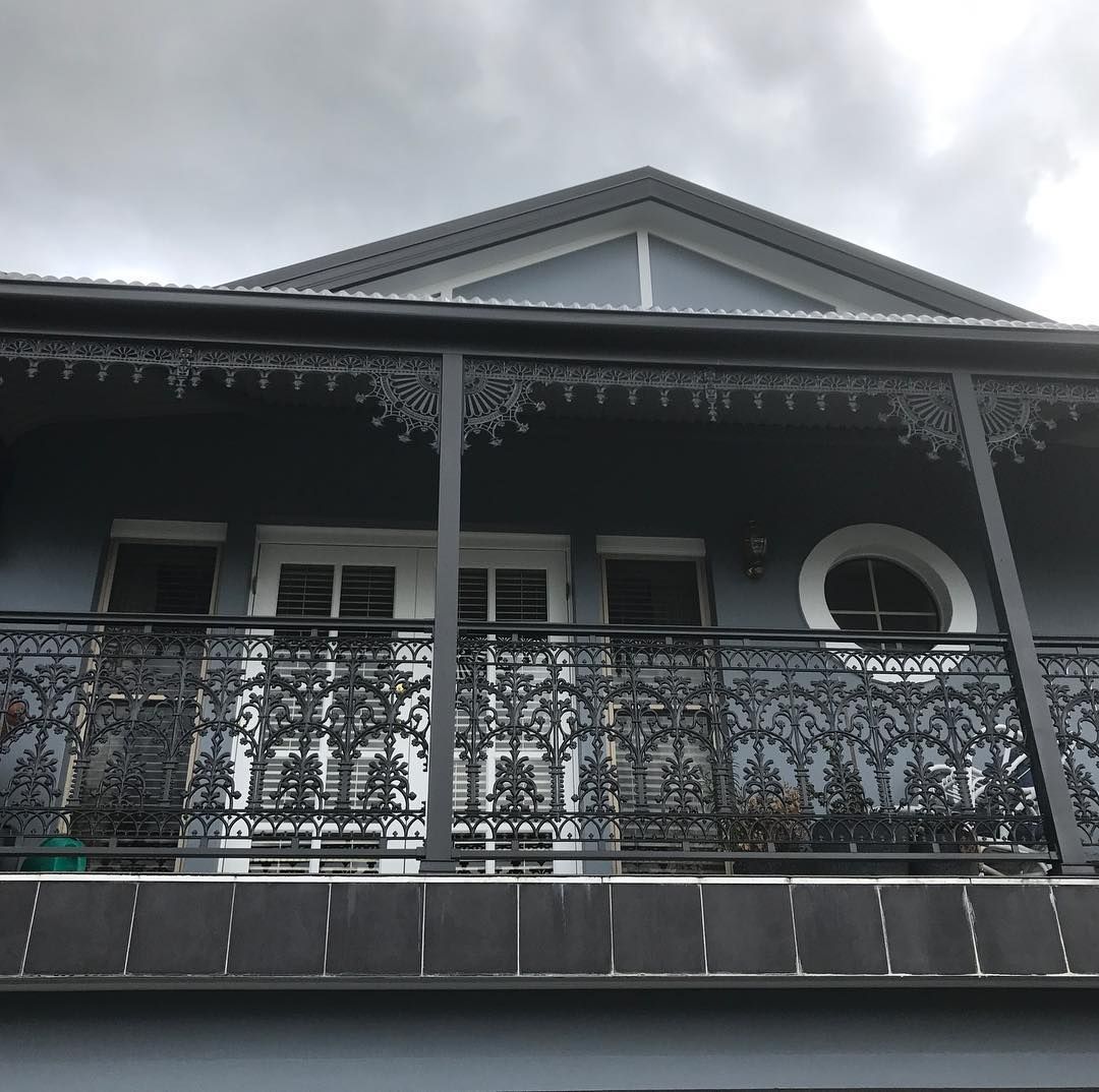 Balcony with ornate black metal railing on a blue building with white shutters. Overcast sky.
