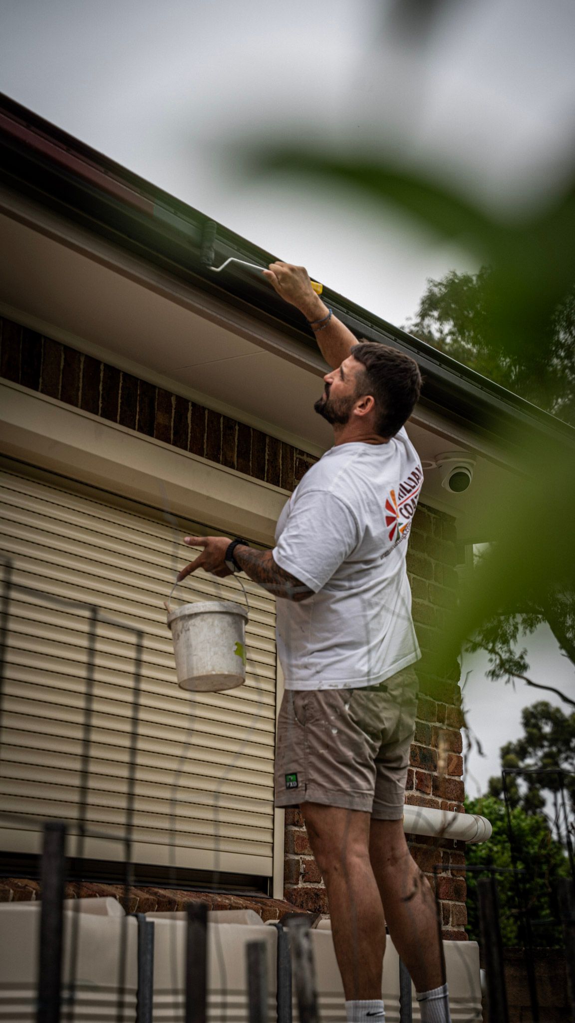 Man on a ladder painting a house gutter, holding a bucket and brush. Outdoors, sunny.