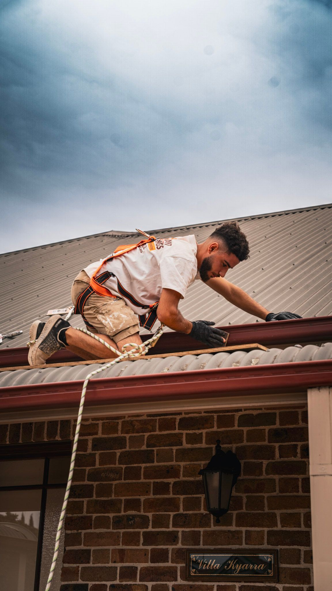 Roofer kneeling on a roof, wearing a harness, working on a gutter. Brick house, cloudy sky.