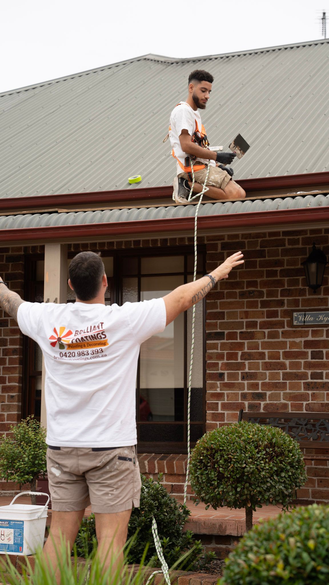 Two men cleaning a house gutter. One man on the roof, using tools. The other man below with arms outstretched.