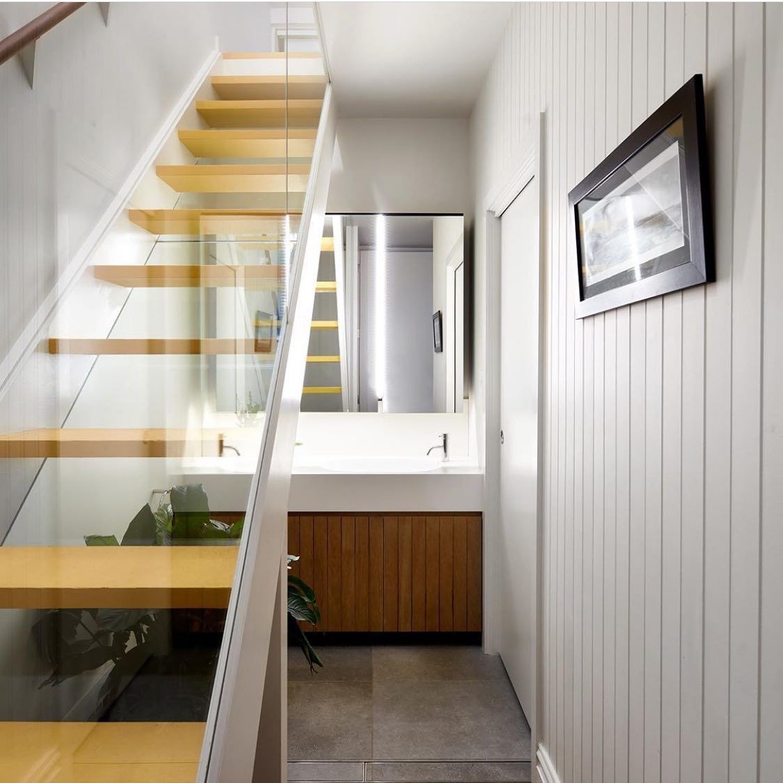 Staircase with glass railing next to a narrow hallway with a sink, mirror, and artwork.