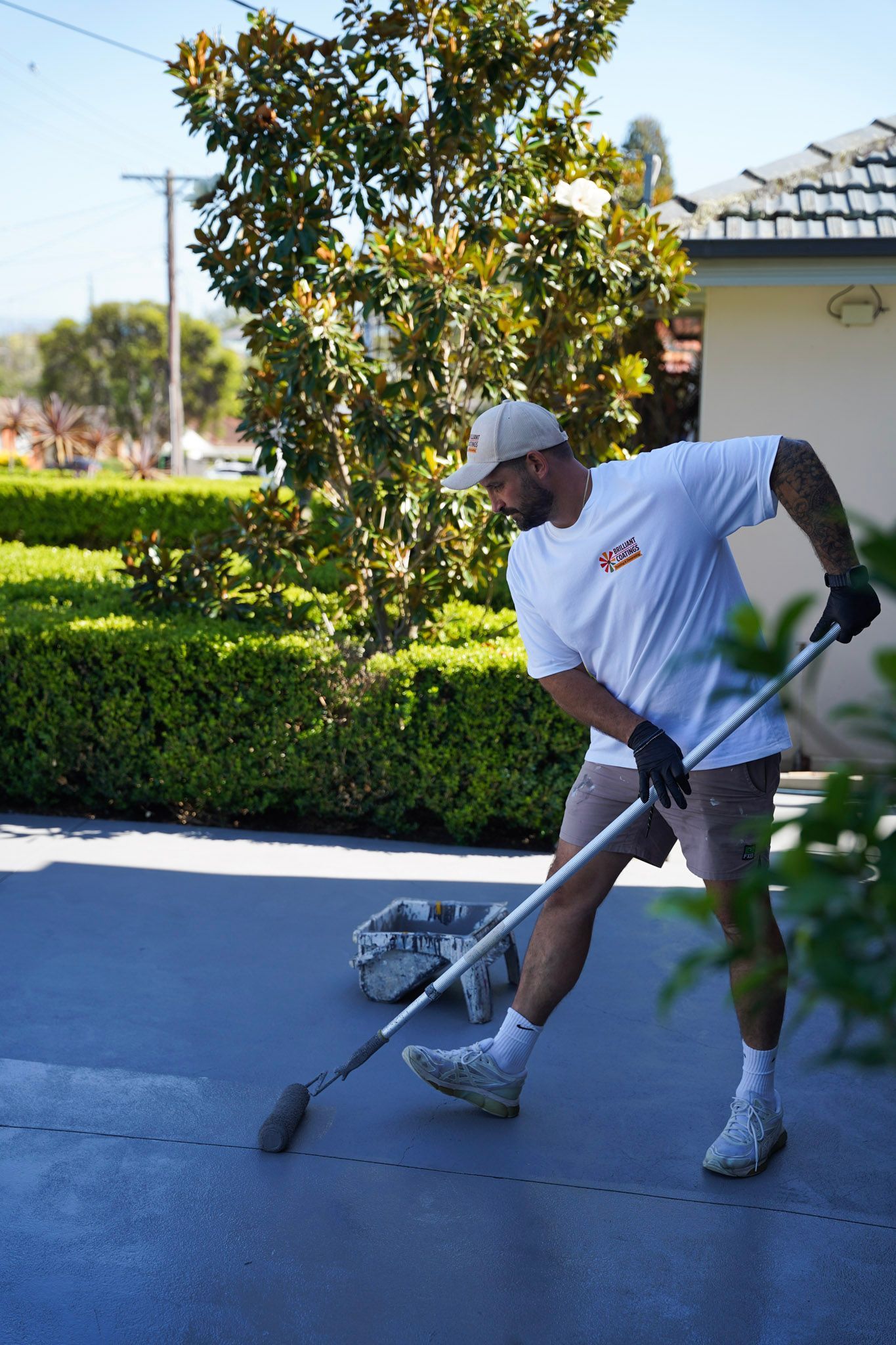 Man in a white shirt and shorts sweeping a dark driveway with a long-handled brush, with a house and greenery in the background.