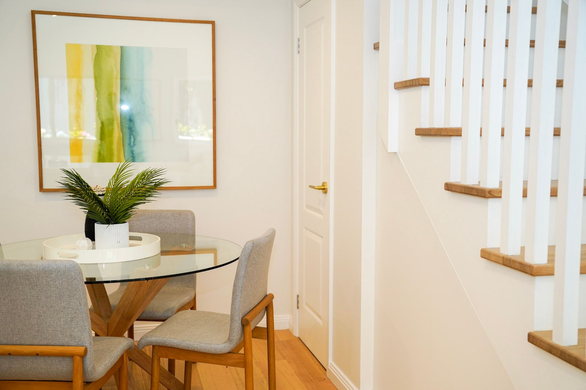 Dining area with glass table, four gray chairs, artwork, staircase with white railings.