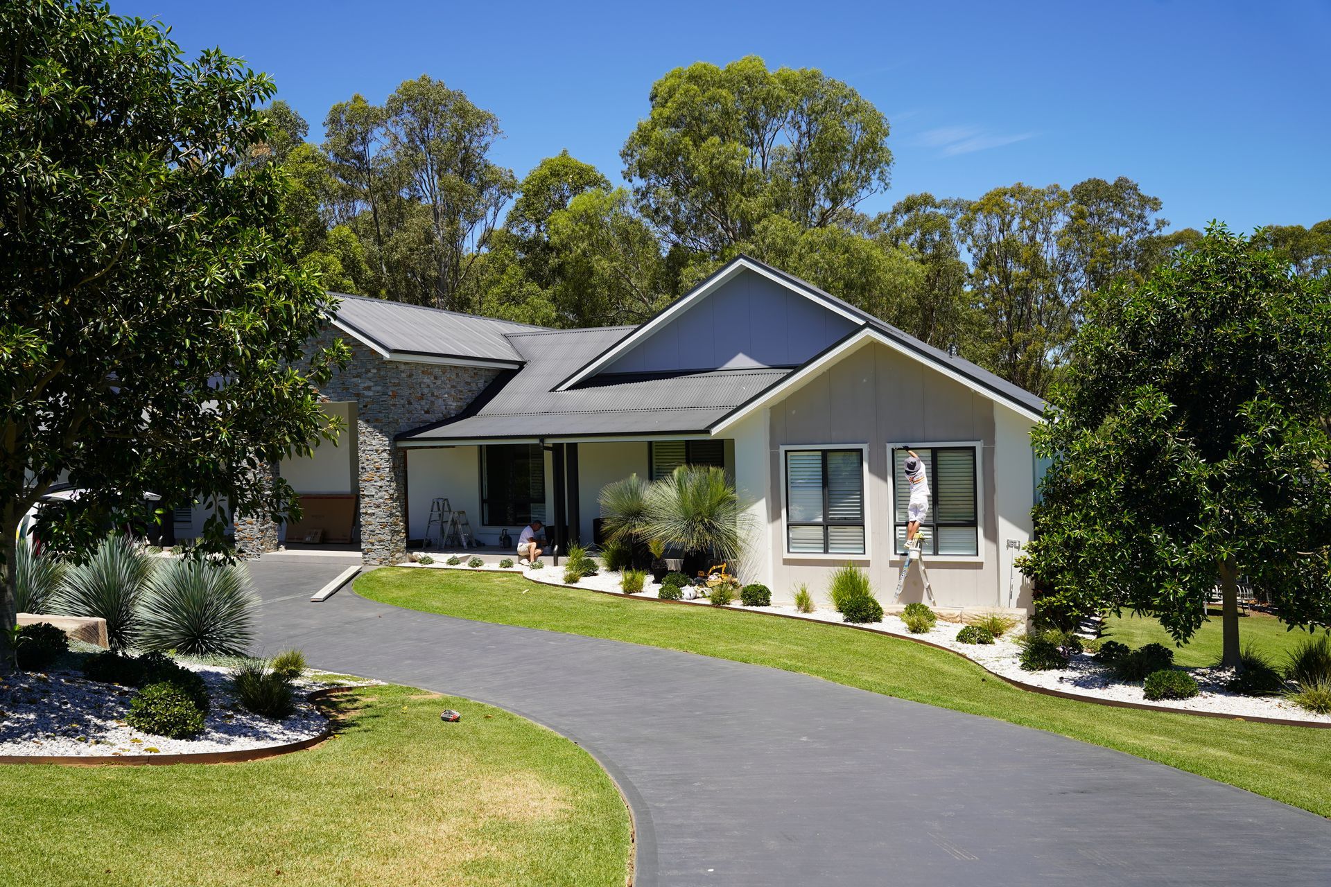 House with a black driveway, light-colored siding, and a dark gray roof, surrounded by trees and green grass.