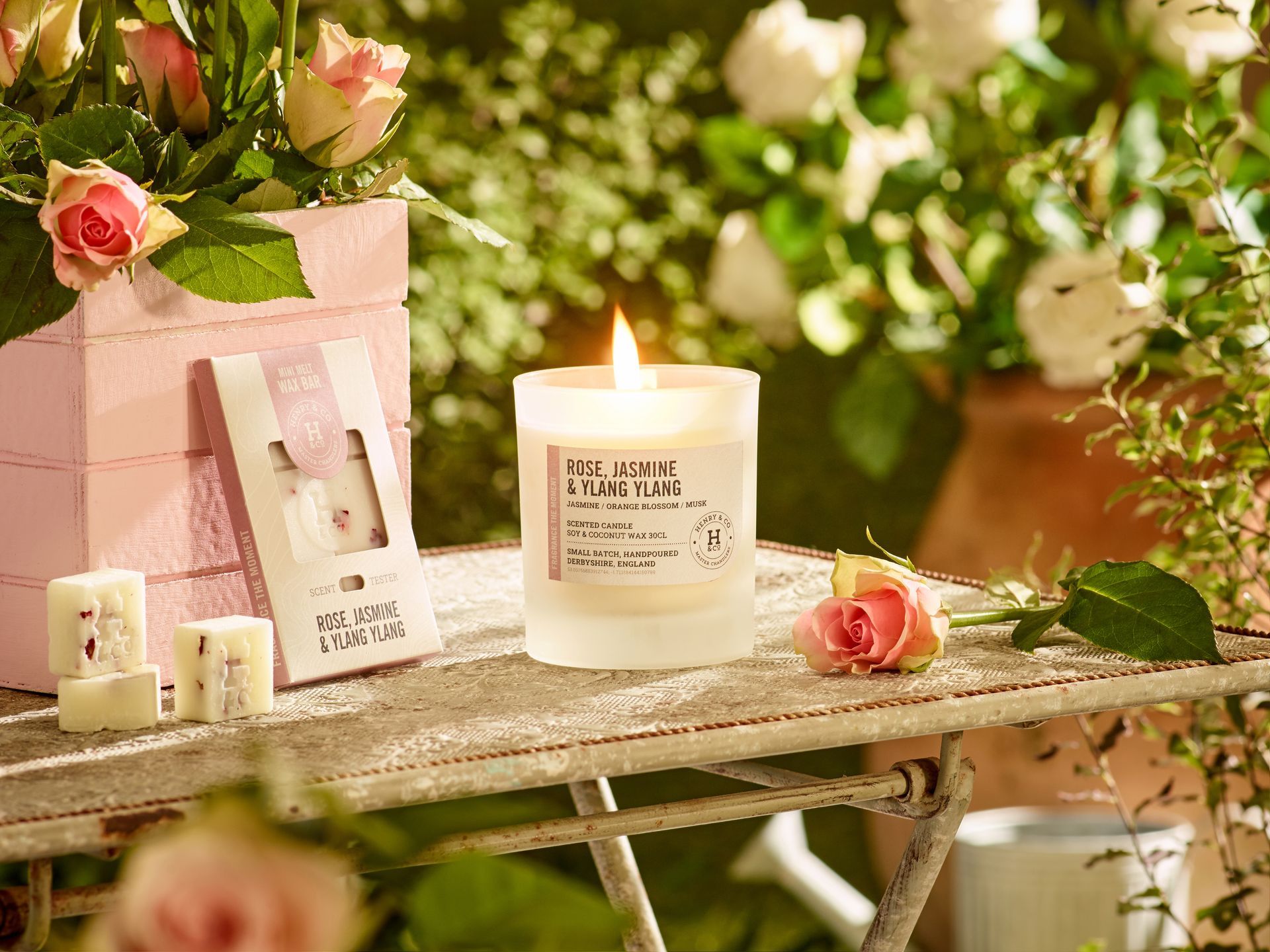Lit candle, flowers, and soap on a rustic table in a garden setting.