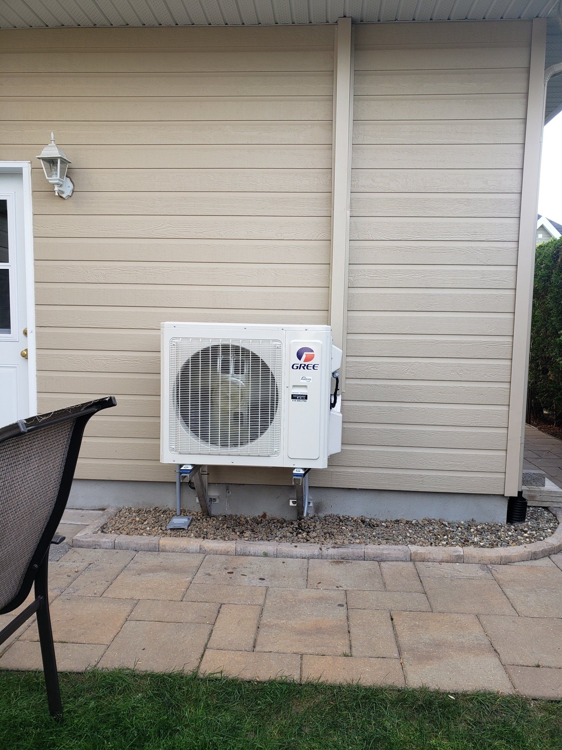 Unité de climatisation extérieure attachée à une maison beige à côté d'un patio en pierre et d'herbe verte.