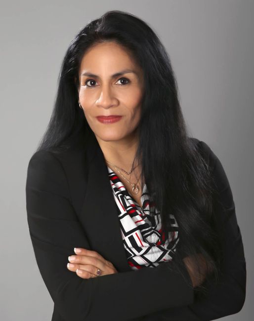 A woman with long black hair and a black jacket is standing with her arms crossed.