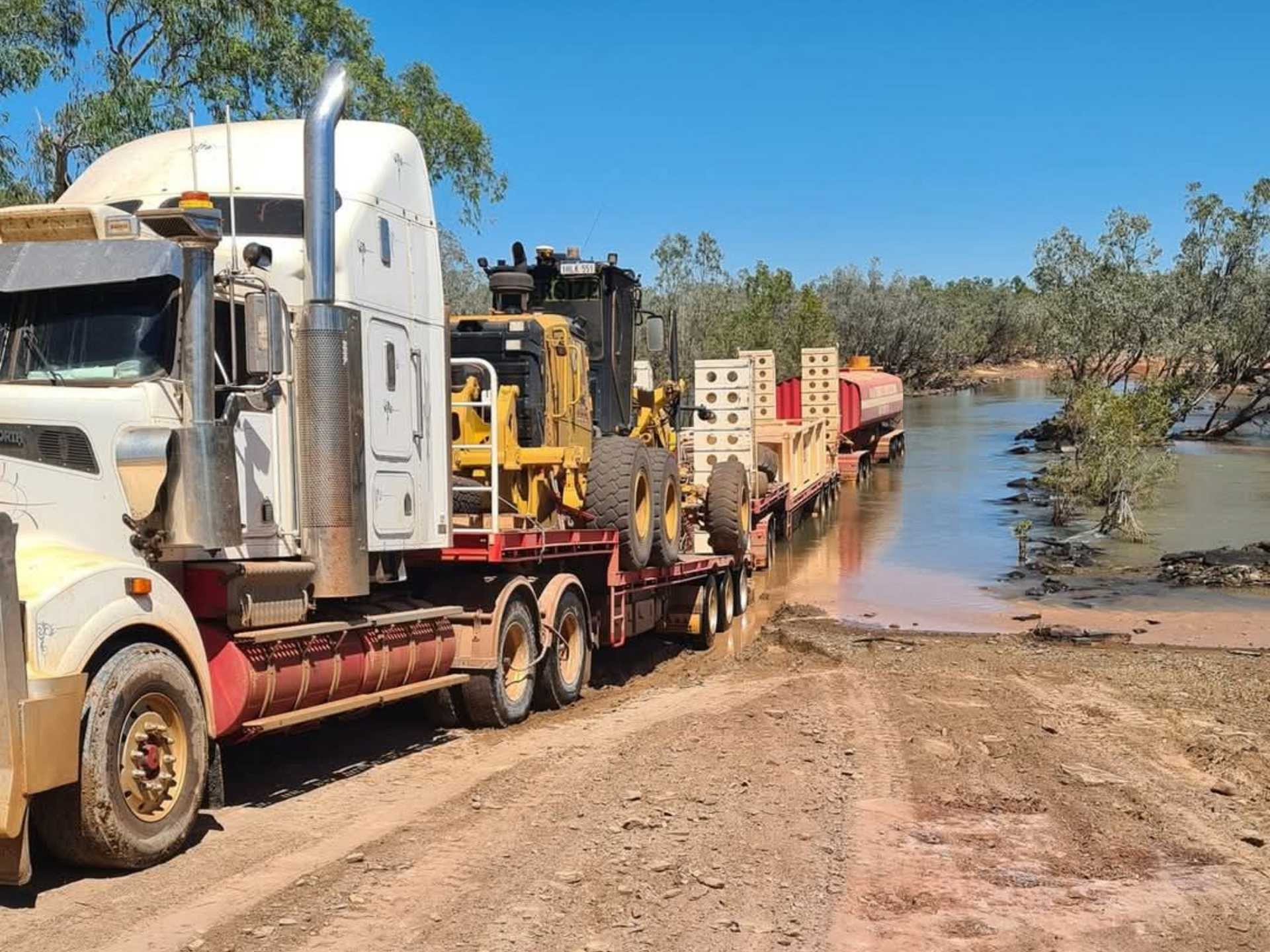 Two yellow road graders working on a dirt road under a clear blue sky.